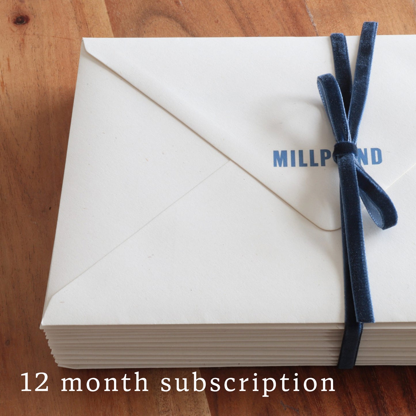 Stack of Millpond envelopes with a blue ribbon on a wooden surface, labeled '12 month subscription'.
