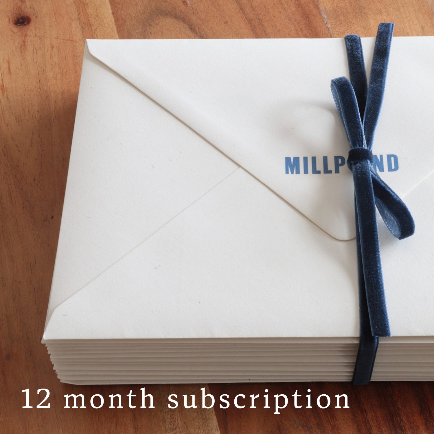 Stack of Millpond envelopes with a blue ribbon on a wooden surface, labeled '12 month subscription'.